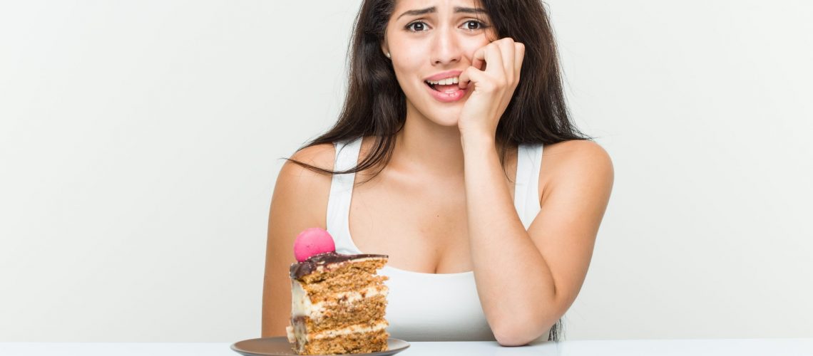 Young hispanic woman eating a cake biting fingernails, nervous and very anxious.
